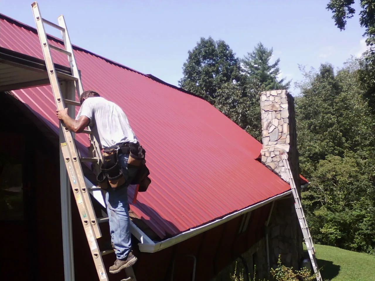 Roofer on a ladder beside a red metal roof and stone chimney — Soffit & Fascia Repair by Roofing Brothers in Willows